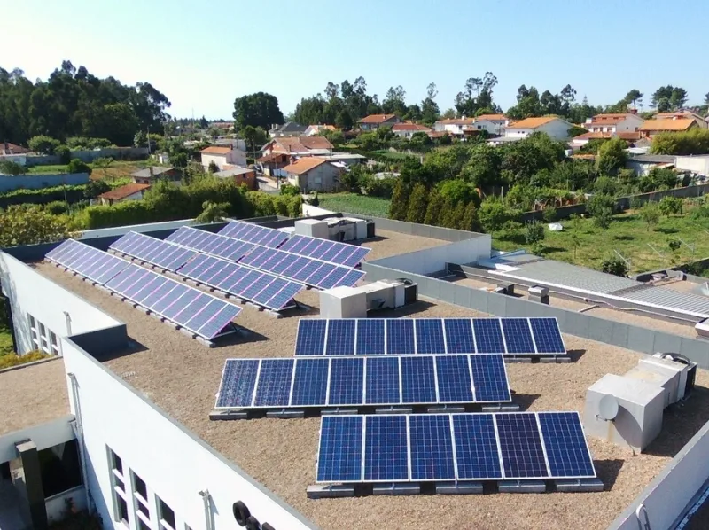 Painéis solares na cobertura plana de um edifício, com casas e árvores ao fundo